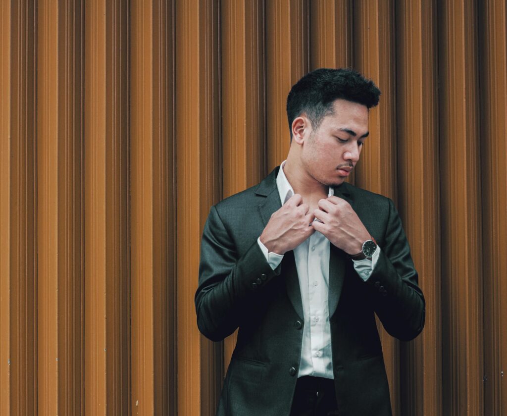 A fashionable young man in a suit adjusts his jacket against a textured wall, exuding confidence and style.